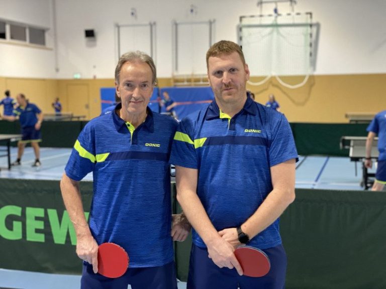 Zwei Tischtennisspieler in blauen Trikots stehen auf einem Tischtennisplatz.
